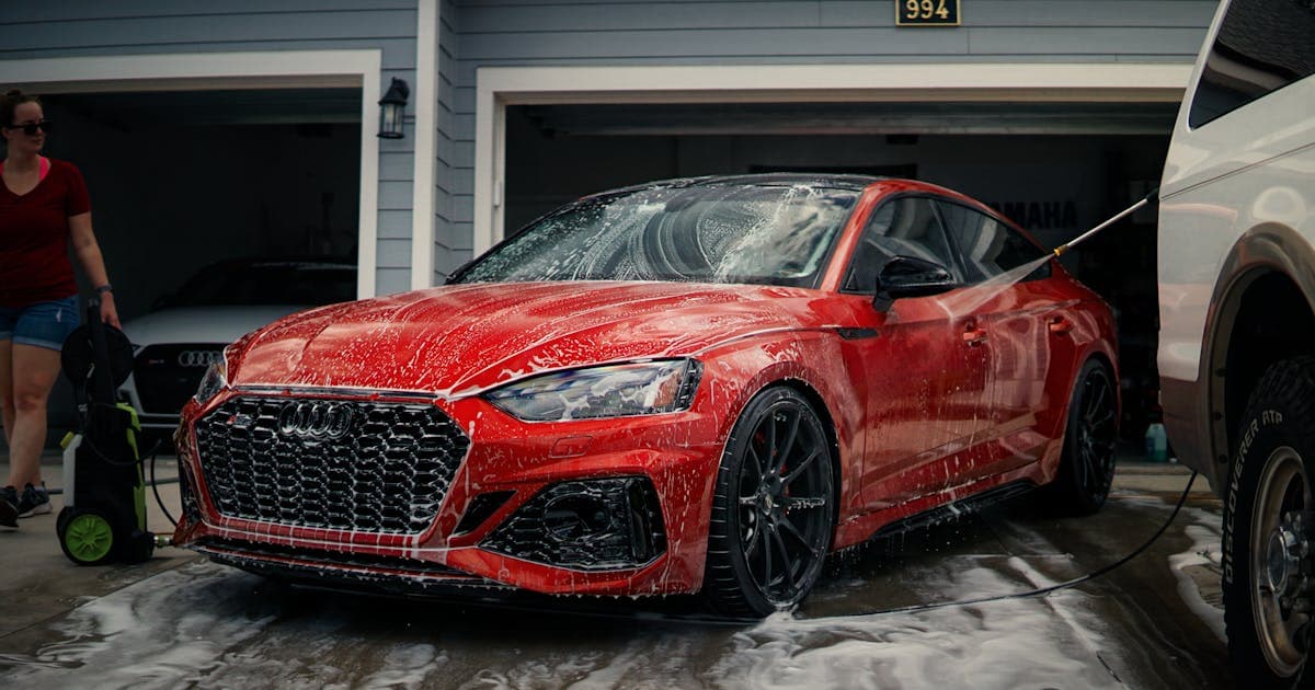 Red sports car being washed with soap in a driveway with a modern garage in the background
